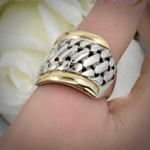 May include: A wide band ring featuring a central woven silver-tone mesh pattern flanked by smooth, polished gold-tone borders on the top and bottom edges. The design creates a textured contrast between the metallic silver center and the gold bands.