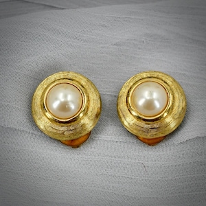 May include: A pair of round clip-on earrings featuring a central cream-colored faux pearl set within a textured gold-tone metal frame. The circular gold border displays fine, concentric etched lines surrounding the smooth, glossy pearl center.