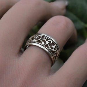 Può includere: Un anello in metallo color argento caratterizzato da una fascia larga con un intricato motivo in filigrana traforata di linee curve interconnesse. Il design è delimitato da bordi sottili e testurizzati che incorniciano il dettaglio.