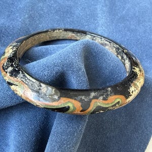 Puede incluir: Un brazalete negro con un patrón decorativo en naranja, verde y dorado. El brazalete tiene una apariencia ligeramente desgastada, lo que sugiere edad o un estilo vintage. El brazalete es circular y parece estar hecho de un material sólido.