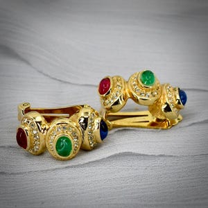 May include: A pair of gold-tone clip-on earrings featuring three oval cabochon stones in red, green, and blue. Each stone is surrounded by a halo of small clear crystals set into the metal frame, creating a multi-colored, textured appearance.