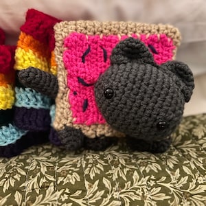 Puede incluir: Un juguete de peluche de ganchillo con forma de gato que sostiene un cuadrado de ganchillo rosa y marrón con una cara sonriente. El gato es gris y tiene ojos negros.