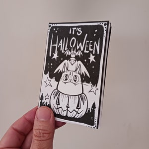 May include: A black and white illustration of a cat wearing a bat costume and sitting on top of a jack-o'-lantern. The text "IT'S HALLOWEEN" is written above the cat.