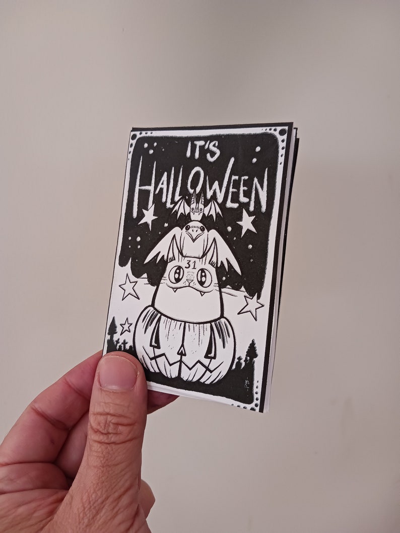 May include: A black and white illustration of a cat wearing a bat costume and sitting on top of a jack-o'-lantern. The text "IT'S HALLOWEEN" is written above the cat.