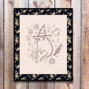 May include: A framed art print featuring a whimsical illustration of a cat wearing a witch's hat, surrounded by magical symbols like stars, crystals, and a bird. The frame has a dark floral pattern.