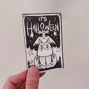 May include: Black and white illustration of a cat wearing a pumpkin costume with a bat on its head. The text "It's Halloween" is written above the cat.