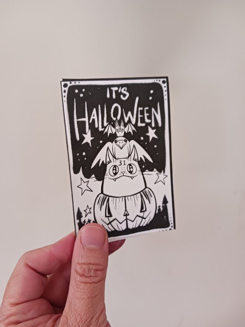 May include: Black and white illustration of a cat wearing a pumpkin costume with a bat on its head. The text "It's Halloween" is written above the cat.