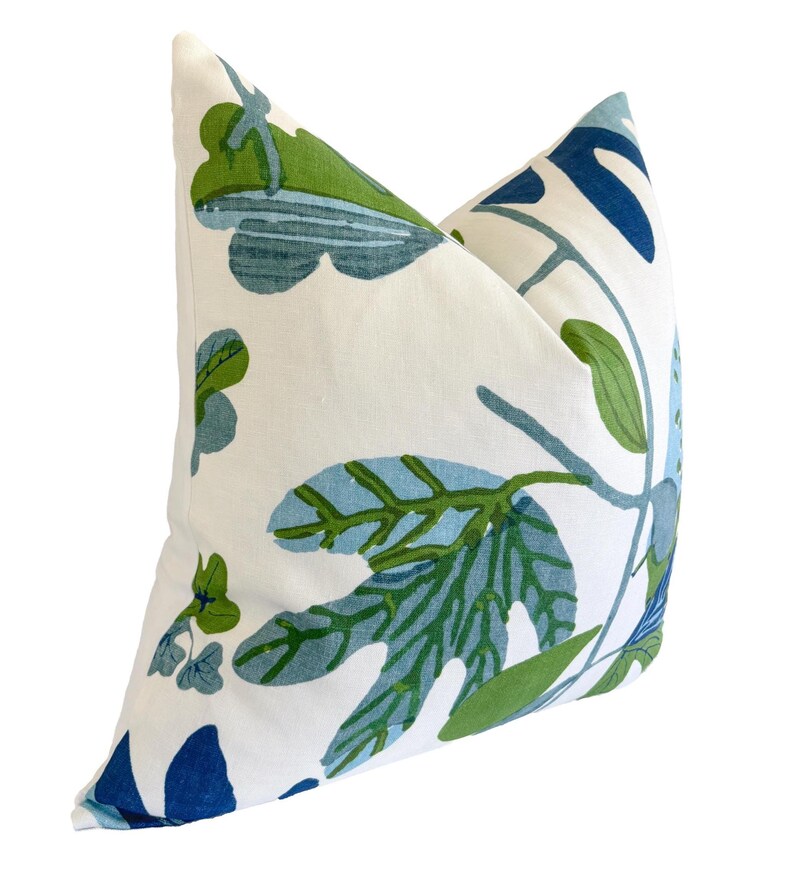 May include: A square cushion with a white base, adorned with a botanical design. The pattern showcases large leaves and stems in shades of blue and green. The cushion has a soft appearance, suitable for a living space.