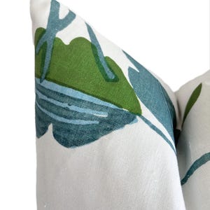 May include: Close-up of a decorative cushion with a white backdrop. The cushion displays a repeating pattern of green and teal tropical leaves. The leaves are stylized, creating a bold, modern design. The cushion is made of a textured fabric.