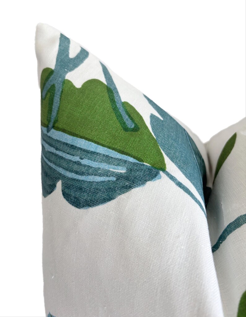 May include: Close-up of a decorative cushion with a white backdrop. The cushion displays a repeating pattern of green and teal tropical leaves. The leaves are stylized, creating a bold, modern design. The cushion is made of a textured fabric.