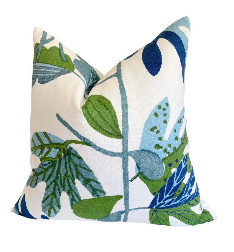 May include: Square cushion with a white backdrop and a botanical design of green, blue, and navy leaves and stems. The cushion has a soft, textured look and is likely made of fabric.