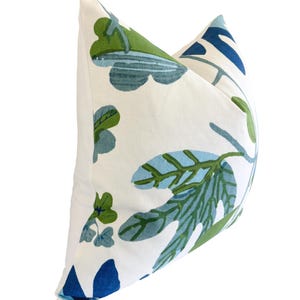 May include: A cushion featuring a white backdrop adorned with a repeating pattern of green and blue leaves and floral motifs. The cushion has a soft, textured look, suitable for home decoration.