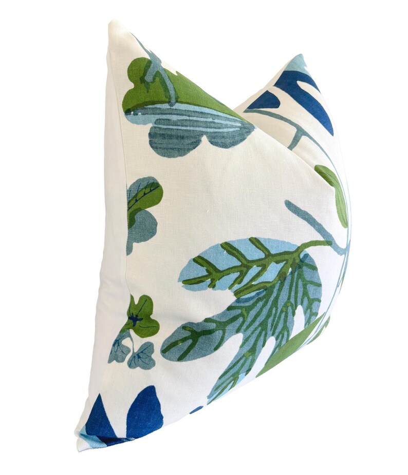 May include: A cushion featuring a white backdrop adorned with a repeating pattern of green and blue leaves and floral motifs. The cushion has a soft, textured look, suitable for home decoration.