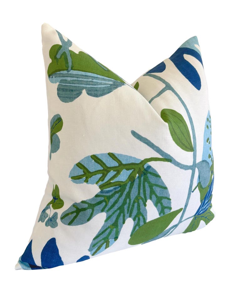 May include: A square cushion with a white base, adorned with a botanical design. The pattern showcases large leaves and stems in shades of blue and green. The cushion has a soft, inviting appearance, perfect for a sofa or bed.
