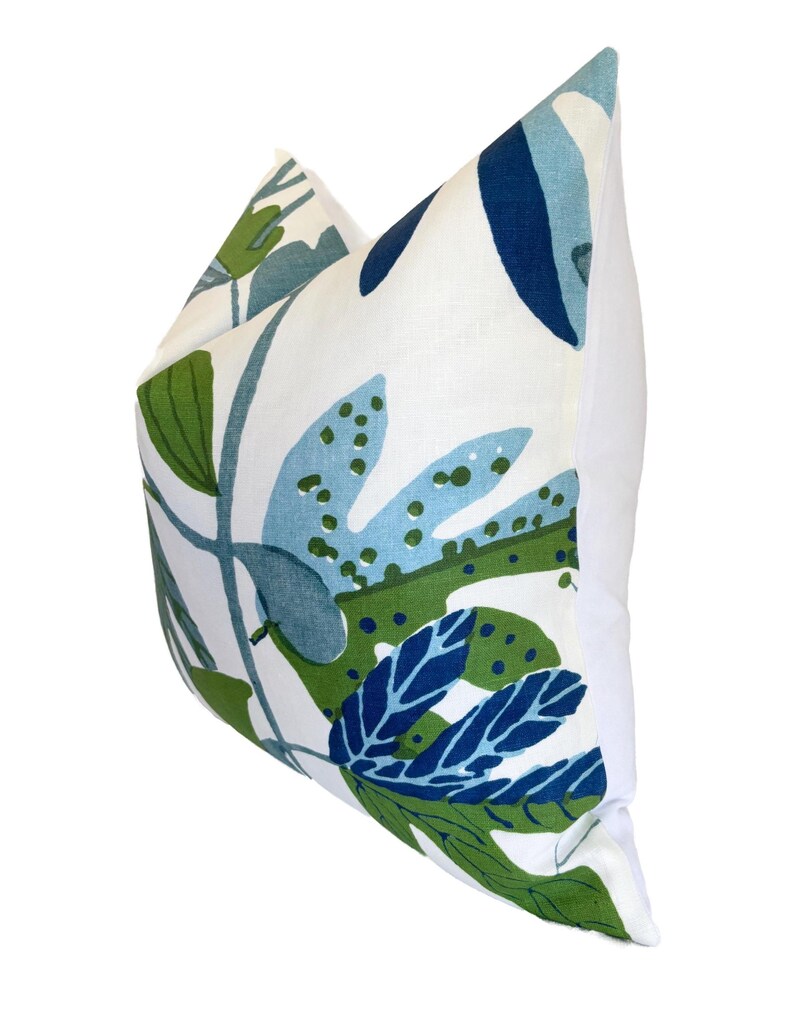 May include: A cushion with a white background and a botanical design. The pattern includes large leaves in shades of blue and green. The cushion has a plain white reverse.