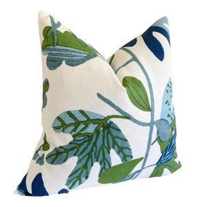 May include: A square cushion with a white base and a botanical design. The pattern includes green and blue leaves and stems. The cushion is likely made of fabric and is designed for decorative purposes.