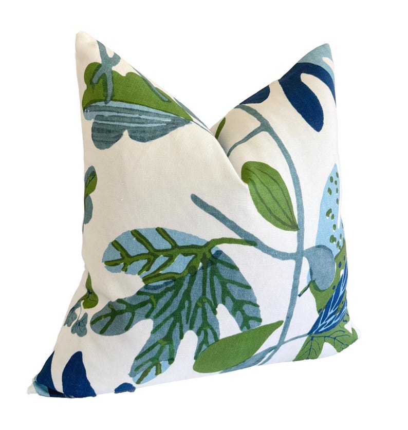 May include: A square cushion with a white base and a botanical design. The pattern includes green and blue leaves and stems. The cushion is likely made of fabric and is designed for decorative purposes.