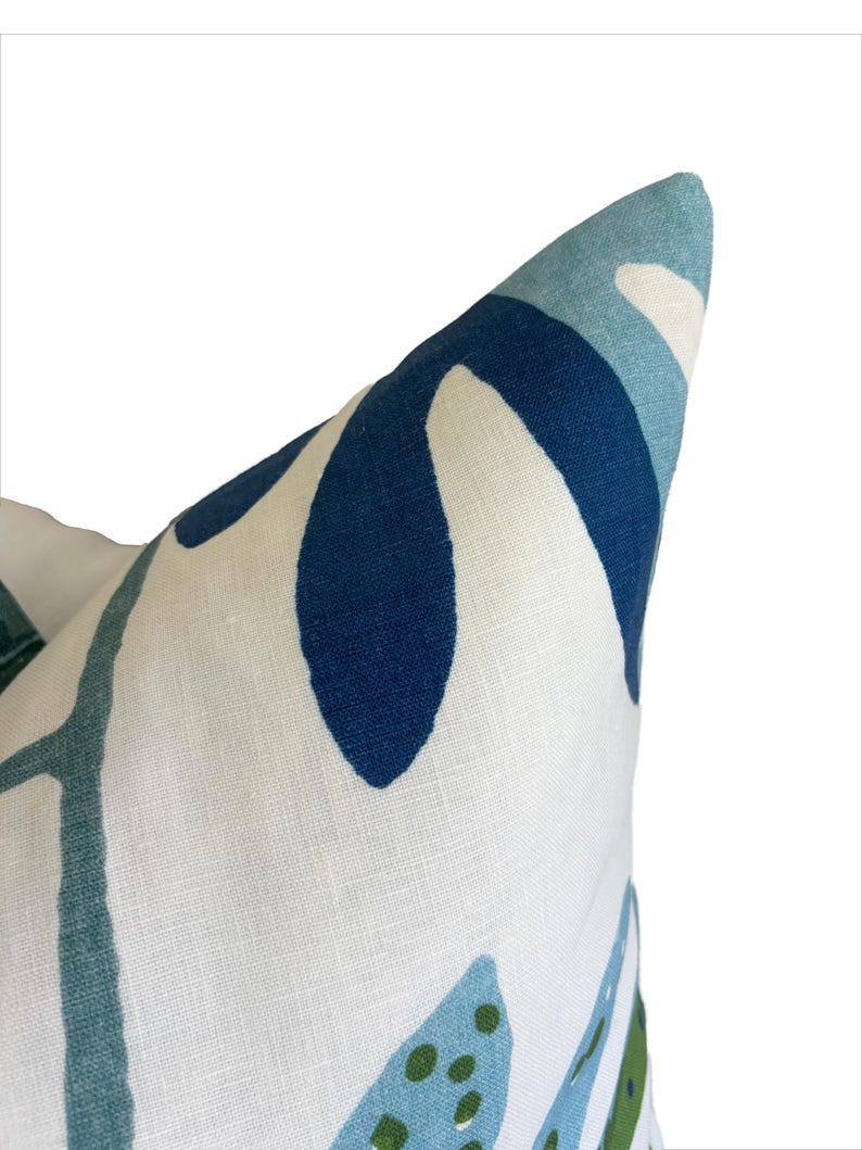 May include: Close-up of a decorative cushion with a white background. The cushion features a pattern of blue and green leaf-like shapes on a white linen fabric. The design includes shades of navy, teal, and light blue, creating a fresh, modern aesthetic.