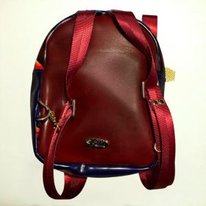 May include: Burgundy leather backpack with red straps and blue trim. The backpack has a silver metal clasp and a small gold tag. The straps are adjustable and the bag appears to be new.