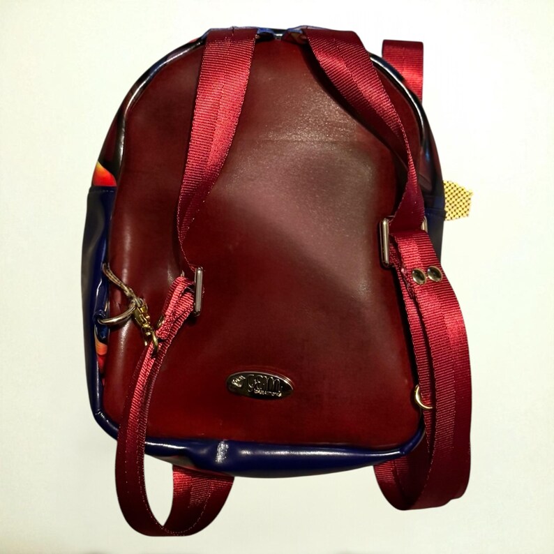 May include: Burgundy leather backpack with red straps and blue trim. The backpack has a silver metal clasp and a small gold tag. The straps are adjustable and the bag appears to be new.