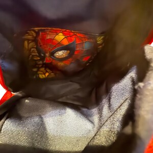 May include: A close-up of a Spider-Man mask with a red, black, yellow, and orange color scheme. The mask is partially visible inside a black bag with red trim. The mask has a webbed design.