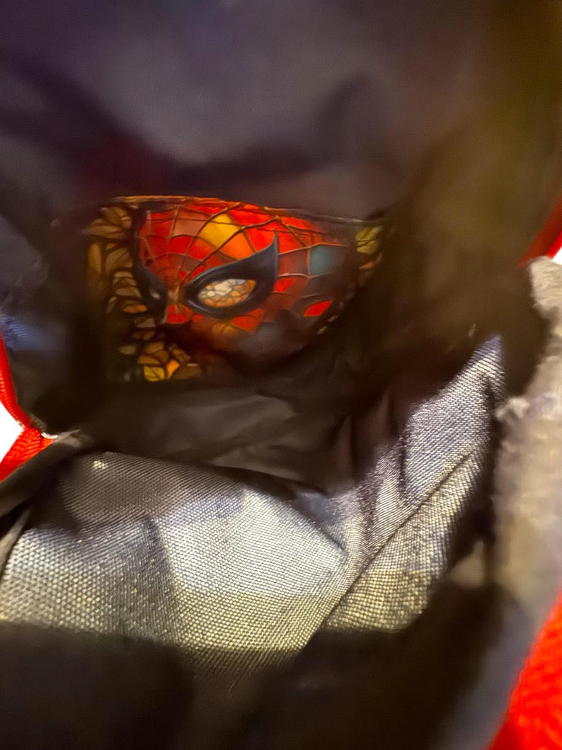May include: A close-up of a Spider-Man mask with a red, black, yellow, and orange color scheme. The mask is partially visible inside a black bag with red trim. The mask has a webbed design.