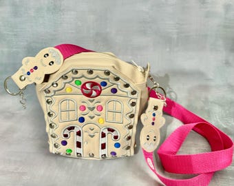 May include: A beige gingerbread house-shaped crossbody bag with a pink strap and gingerbread man charms. The bag is decorated with colorful candy and candy cane accents.