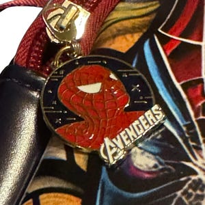 May include: Close-up of a black and red bag with a red zipper and a gold zipper pull. A circular charm features a red Spider-Man head and the word "AVENGERS" in silver.