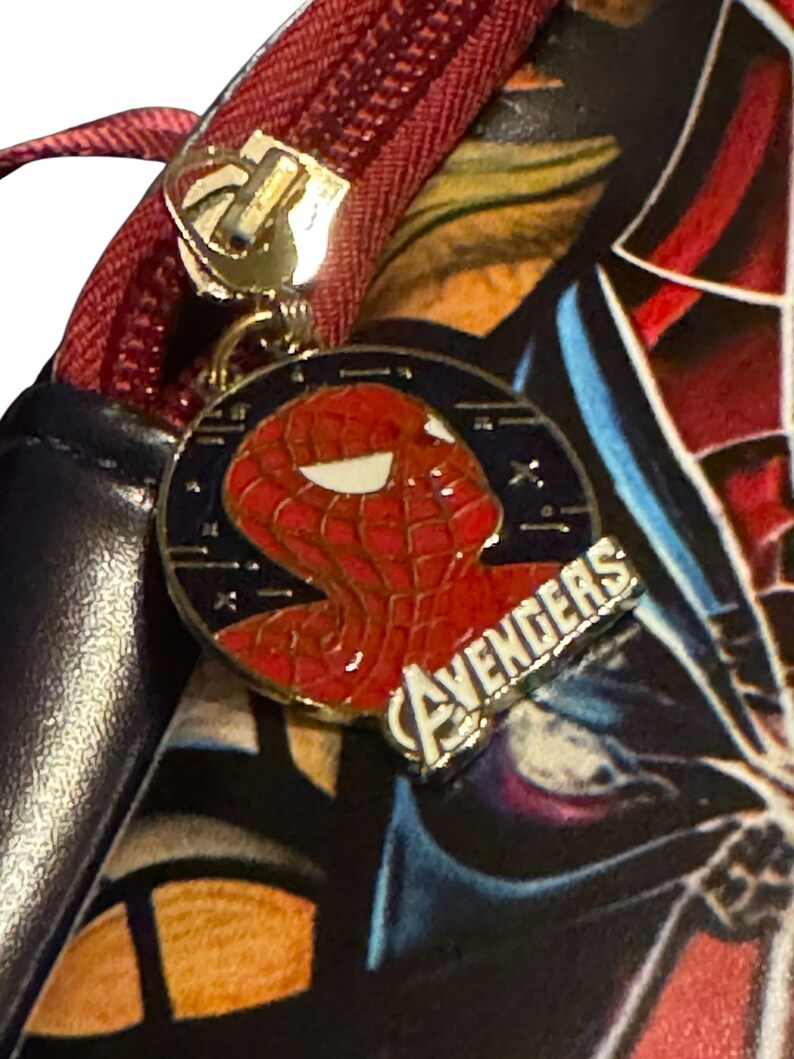 May include: Close-up of a black and red bag with a red zipper and a gold zipper pull. A circular charm features a red Spider-Man head and the word "AVENGERS" in silver.