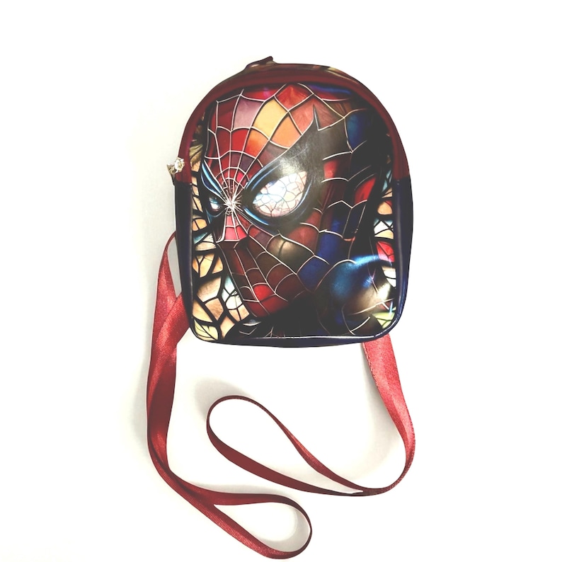 May include: A small, burgundy and navy blue backpack featuring a Spiderman graphic on the front. The graphic shows Spiderman's face in vibrant colors. The bag has a burgundy shoulder strap and a zipper closure.