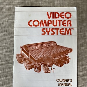 Pode incluir: Um manual do usuário branco do Atari Video Computer System com um logotipo vermelho da Atari e uma imagem do console e dos controladores. O manual é para o modelo CX2600A.