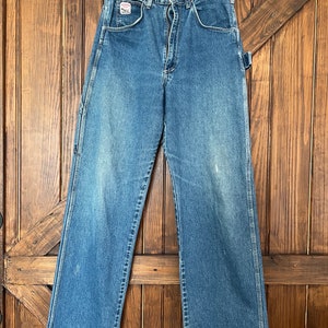 May include: Blue denim work pants with a straight leg, hanging on a wooden hanger. The pants have a classic five-pocket design, a zipper fly, and a small logo patch on the front pocket. The pants are made of a durable denim material.