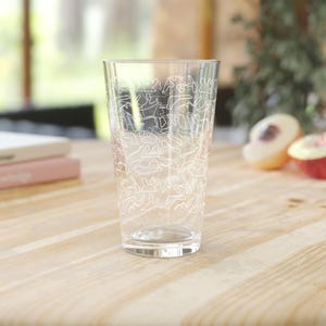 May include: A clear pint glass with a white line drawing pattern of various figures. The glass is cylindrical and sits on a light-colored wooden surface. The background includes books and fruit.
