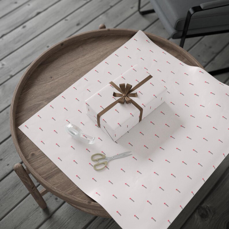 Pu&ograve; includere: Un pacco regalo avvolto con un nastro e un fiocco marrone &egrave; appoggiato su carta da regalo bianca con un motivo rosso ripetuto. Un paio di forbici dorate e un rotolo di nastro adesivo trasparente sono anch'essi sulla carta.