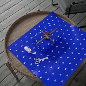 Pu&ograve; includere: Un regalo avvolto con un nastro marrone &egrave; appoggiato su carta da regalo blu con un motivo bianco. Sul foglio sono presenti anche un rotolo di nastro adesivo e delle forbici. Gli oggetti sono su un tavolo di legno.