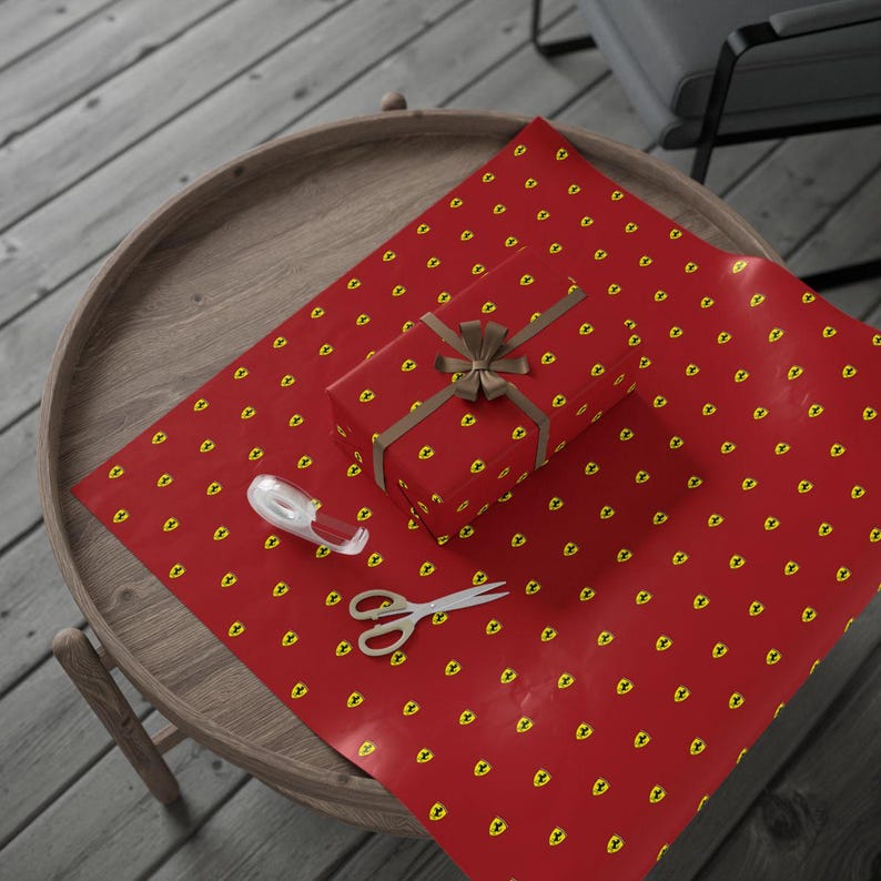 Pu&ograve; includere: Un regalo avvolto su un tavolo di legno. La carta da regalo rossa presenta un motivo ripetuto con logo giallo. Un nastro e un fiocco marroni adornano il pacco. Forbici e nastro adesivo sono nelle vicinanze, suggerendo l'incarto regalo.