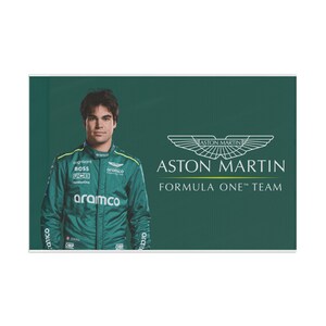 May include: A man wearing a green racing suit with the Aston Martin Formula One Team logo and sponsors' logos. The background is green with the Aston Martin logo and the text "ASTON MARTIN FORMULA ONE™ TEAM".