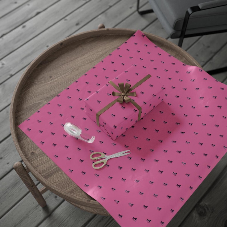 Pu&ograve; includere: Un regalo avvolto su un tavolo rotondo di legno. La carta da regalo rosa presenta un motivo nero ripetuto. Il regalo &egrave; legato con un nastro marrone. Sul tavolo ci sono anche un rotolo di nastro adesivo e delle forbici.