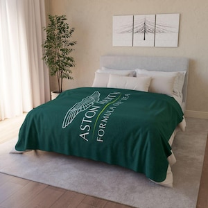 Puede incluir: Una manta verde oscuro con el logotipo y texto de Aston Martin. La manta está sobre una cama con almohadas blancas y un cabecero gris. El texto dice "ASTON MARTIN FORMULA ONE TEAM".