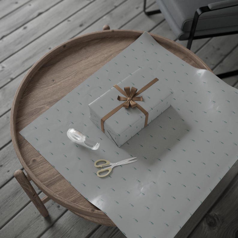 Pu&ograve; includere: Un regalo avvolto con un nastro e un fiocco dorati &egrave; appoggiato su carta da regalo grigia con un motivo sottile. Un rotolo di nastro adesivo e forbici con manici dorati sono anch'essi sulla carta, suggerendo l'incarto.
