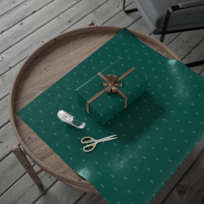 Pu&ograve; includere: Un regalo confezionato su un tavolo rotondo di legno. La carta da regalo &egrave; verde smeraldo con un motivo sottile. Un nastro e un fiocco dorati adornano il pacco. Un dispenser di nastro adesivo trasparente e forbici dorate sono vicini.