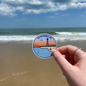 May include: A round sticker featuring a lighthouse scene with the text "Outer Banks, NC". The design includes a sunset, ocean waves, and a black and white lighthouse. The sticker is held up against a beach background.