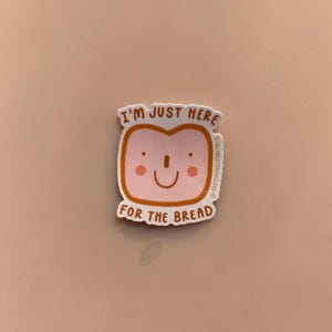 May include: A sticker depicting a cartoon bread slice with a smiling face and pink cheeks. The text "I'M JUST HERE FOR THE BREAD" is printed above and below the bread slice. The sticker is set against a pale pink backdrop.