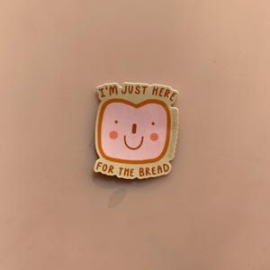 Könnte beinhalten: Ein Aufkleber mit einem lächelnden Brotlaib mit rosa Gesicht und dem Text "I'M JUST HERE FOR THE BREAD" in einer verspielten Schriftart. Der Aufkleber hat einen hellbraunen Rand und ist vor einem rosa Hintergrund platziert.