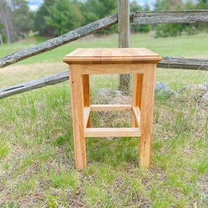May include: A handcrafted wooden stool with a square seat and four sturdy legs. The stool is made of light-colored wood, showcasing the natural grain. A horizontal support bar connects the legs, adding stability. The stool is set on a grassy surface.