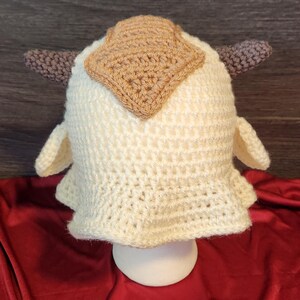 May include: A cream-colored, handmade crochet hat, resembling a character from a popular animated series. The hat features a square patch on top and two small, brown, horn-like projections. The hat is displayed on a white stand against a red background.
