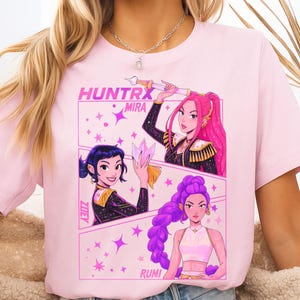 May include: A light pink t-shirt featuring a graphic design with the text "HUNTRX MIRA" and illustrations of three women with colorful hair and outfits. The design includes stars and the names "ZUEY" and "RUMI".