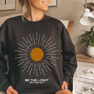 Könnte beinhalten: Schwarzes Sweatshirt mit einem weißen Sonnen-Motiv und dem Text "Sei das Licht Matthäus 5:14".