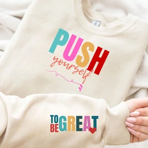 May include: A cream colored sweatshirt with the text "PUSH yourself" in colorful letters on the front and "TO BE GREAT" with a small heart on the sleeve.