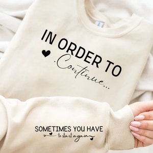 以下が含まれることがあります： ベージュのトレーナーに「In order to continue... Sometimes you have to start again」という黒い文字がプリントされています。テキストには小さな黒いハートが付いています。